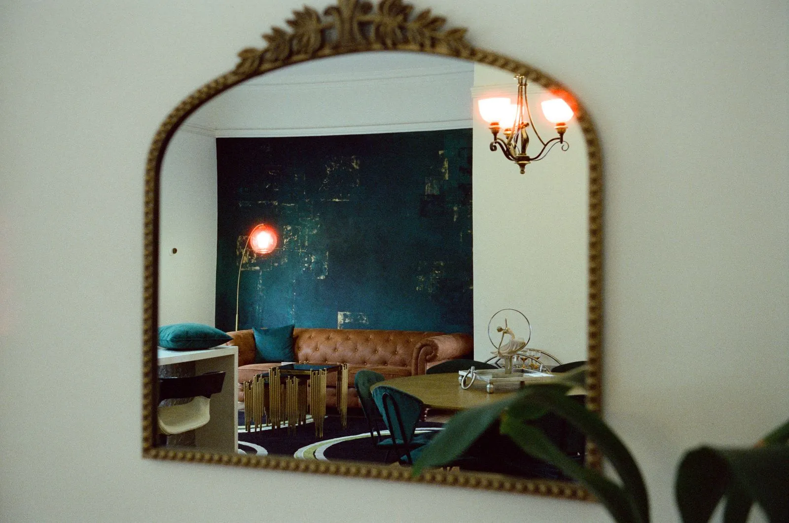 A gilt mirror reflecting a lounge and dining room, chandelier lit above the table
