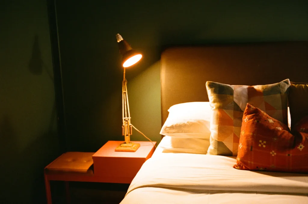 A lamp lit on an orange bedside table beside a made bed