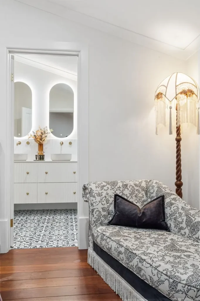 A toile chaise beneath a fringed wall light, the ensuite lit beyond
